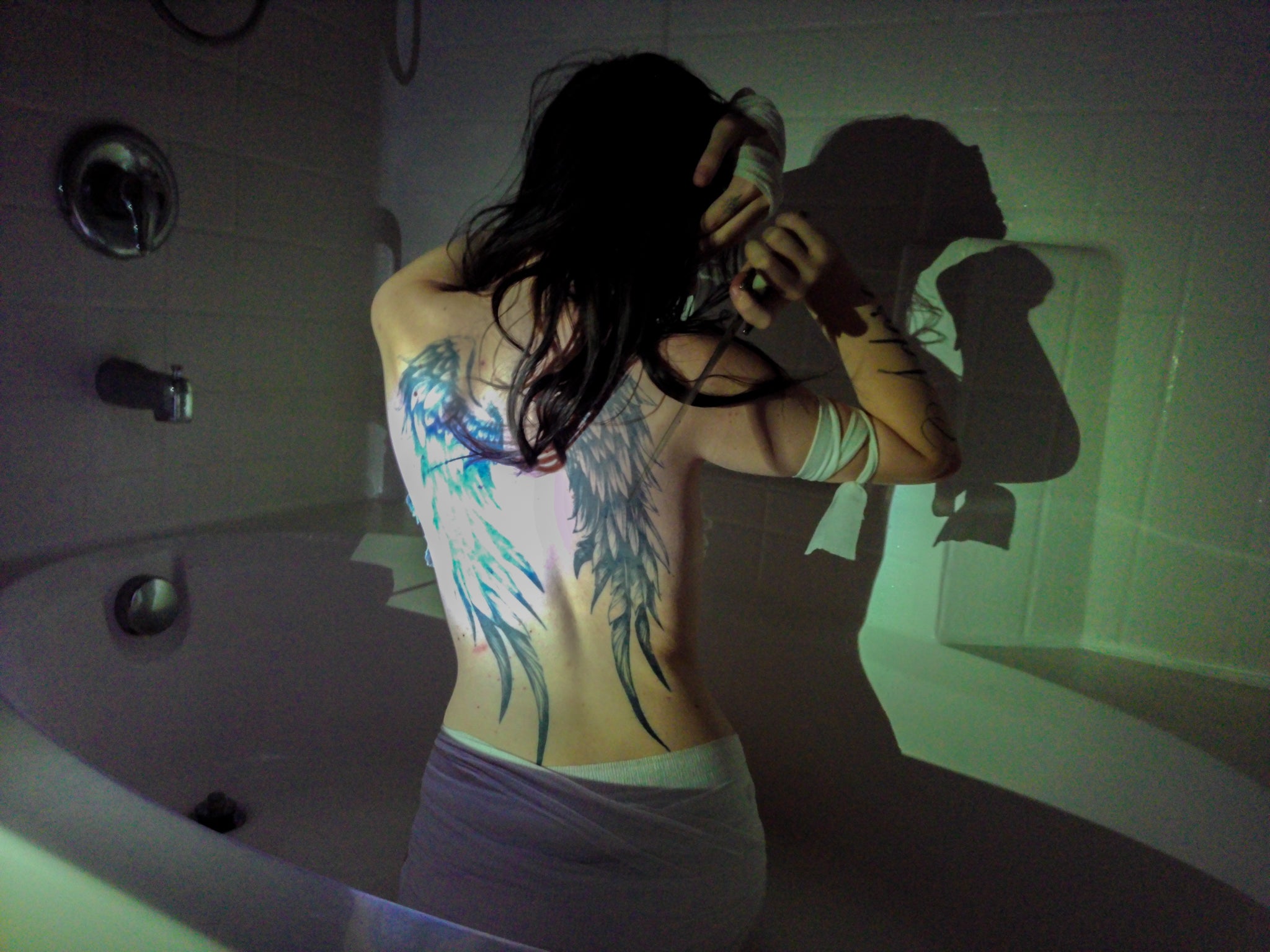 Person with angel wings tattoos in a dimly lit bathroom